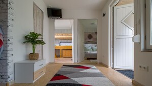Um corredor com piso de madeira, um tapete geométrico, uma planta em vaso, uma TV fixada na parede e uma cama no quarto vizinho.