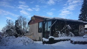 Uma casa com telhado inclinado, antena parabólica e neve no telhado e na vegetação ao redor.