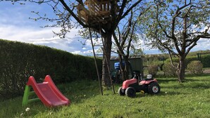Uma área ao ar livre com gramado, uma estrutura de madeira para brincar, um escorrega vermelho e um trator de brinquedo vermelho.