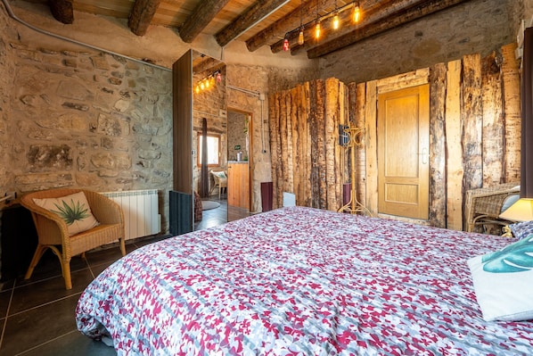 A rustic room with a stone wall, wooden beams, a bed with a floral bedspread, a wicker chair, and a wooden door.