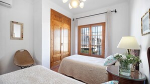 Un dormitorio con dos camas, un armario de madera, una silla, una mesita de noche con lámpara y una ventana con cortinas.