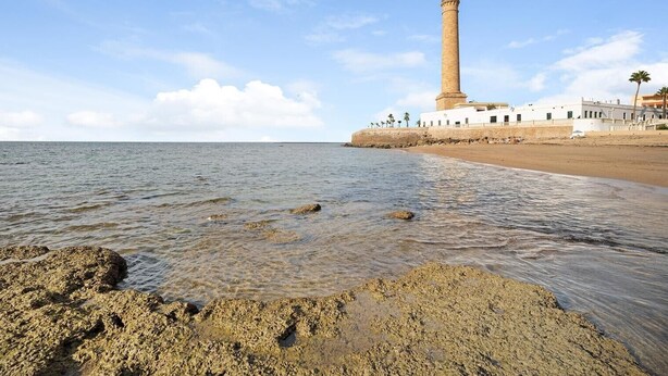 Paisaje costero con un faro, playa arenosa y costa rocosa.