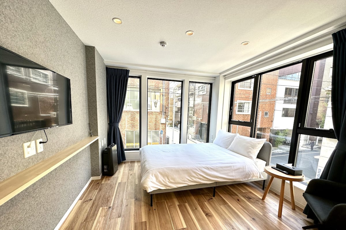 A modern hotel room with a large bed, a TV mounted on the wall, and large windows offering a view of the city.