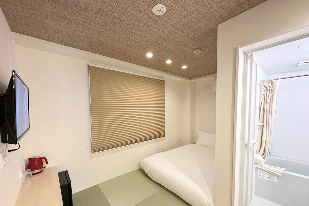 A small, modern room with a bed, a TV mounted on the wall, and a window with blinds.