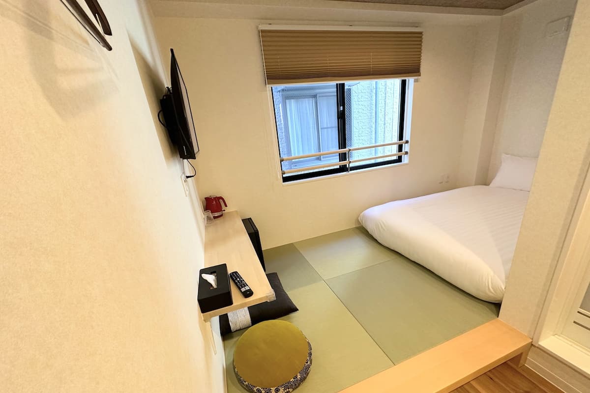 A compact hotel room with a bed, a small table with a remote control, a window with blinds, and a television mounted on the wall.
