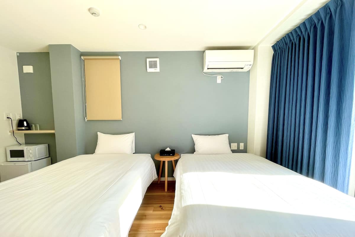 A hotel room with two beds, a microwave, a small table, a wall-mounted air conditioner, and a window with curtains.