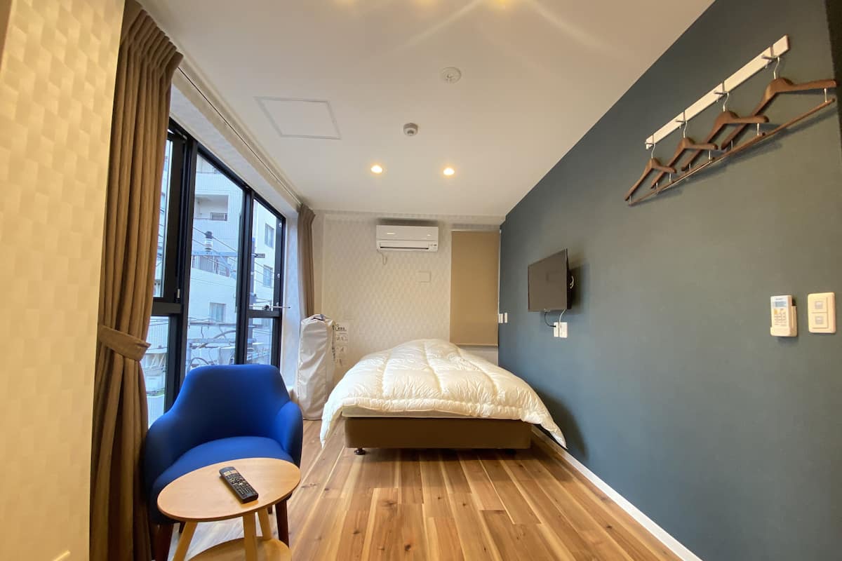 A hotel room with a bed, a blue chair, a small round table, a TV mounted on the wall, and an air conditioning unit.