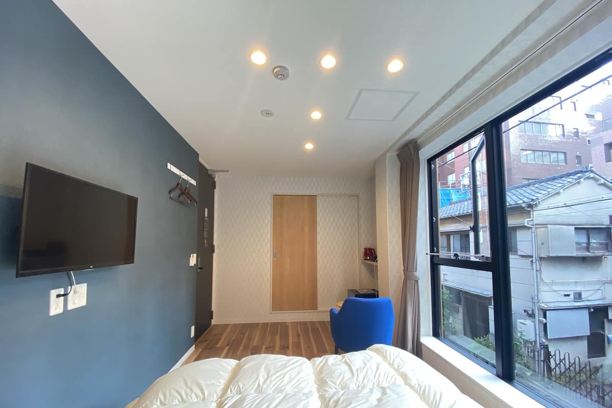 A bedroom with a bed, a TV mounted on the wall, a window with a view of buildings, and a wooden floor.