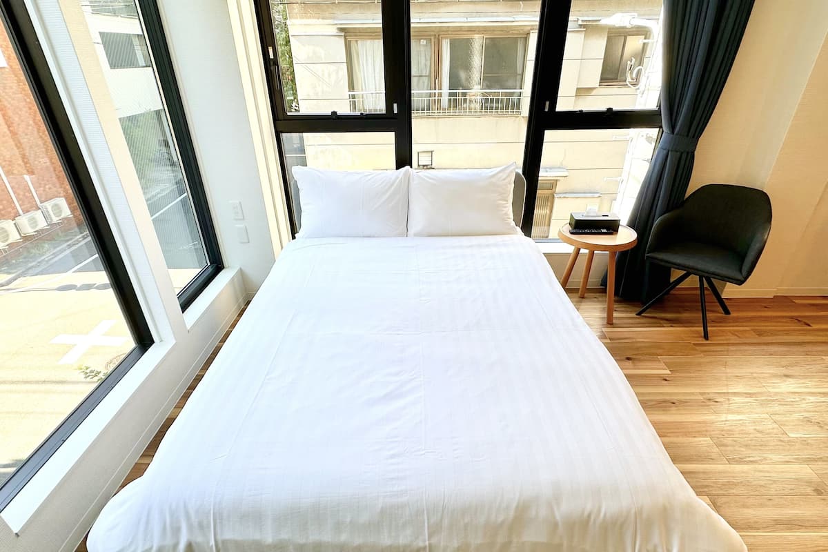 A neatly made bed in a room with large windows, a small round table, and a black chair.