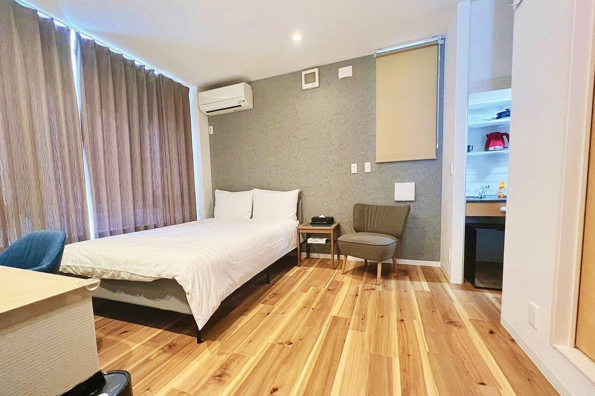 A hotel room with a bed, a desk, a chair, a small table, an air conditioning unit, and a kitchenette.