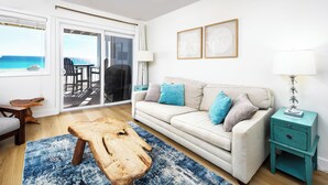 Uma sala de estar com sofá, mesa de centro e vista para o oceano por uma porta de correr.
