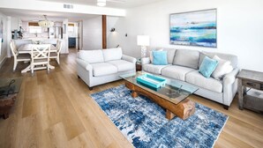 A spacious living room with a glass coffee table, two sofas, a dining area with a chandelier, and a kitchen in the background.