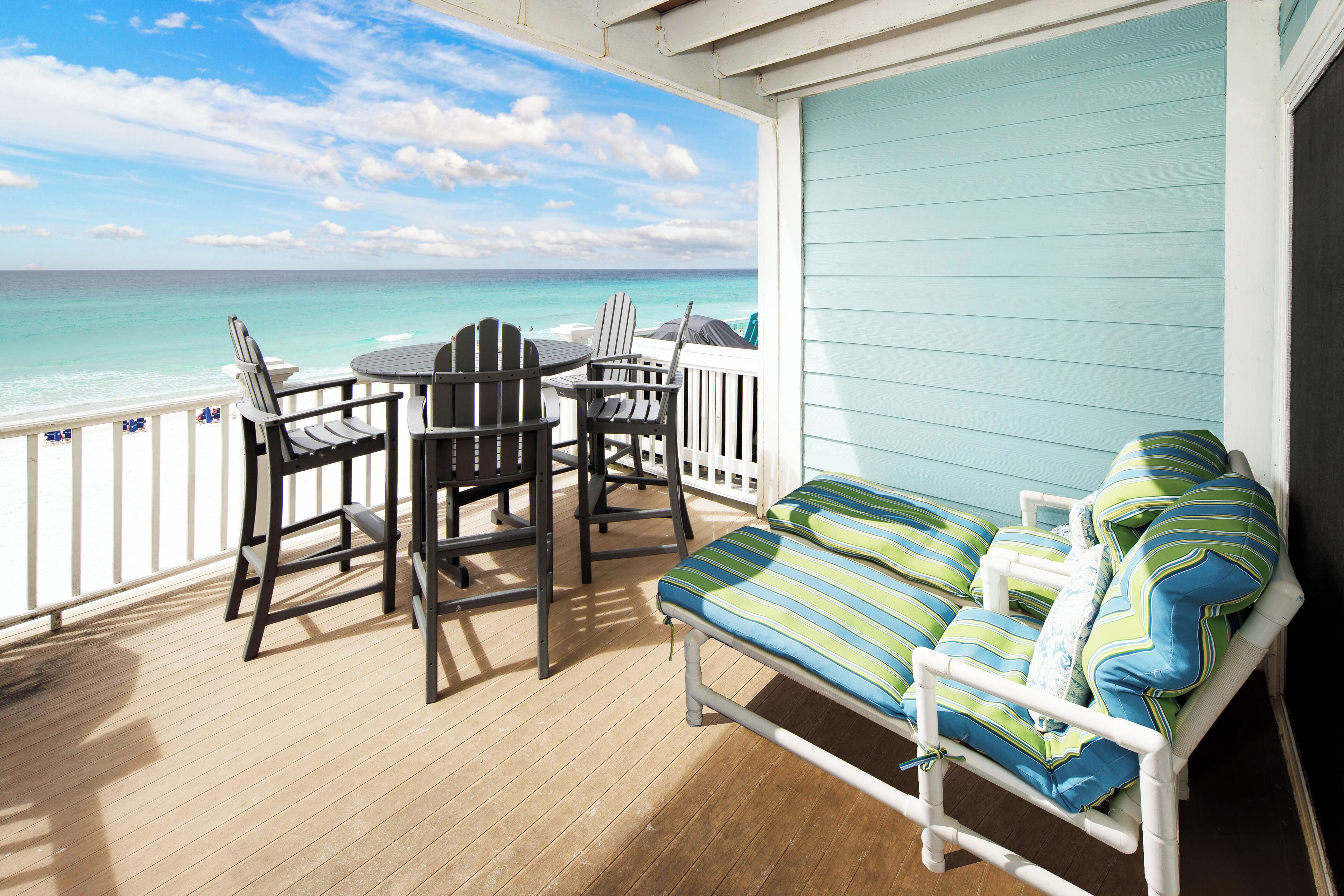 A balcony with a view of the ocean, a white bed with striped pillows, and a table with chairs.