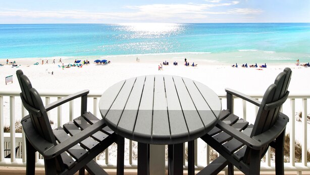 A wooden table and chairs on a balcony overlooking a beach with people and umbrellas.