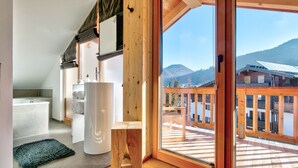 A modern bathroom with a bathtub, a wooden stool, and a large window offering a view of a wooden deck and mountains. - Gipfelhütte - mit Masterschlafzimmer, Zwei Hüttenzimmer und Balkon (Reit im Winkl)
