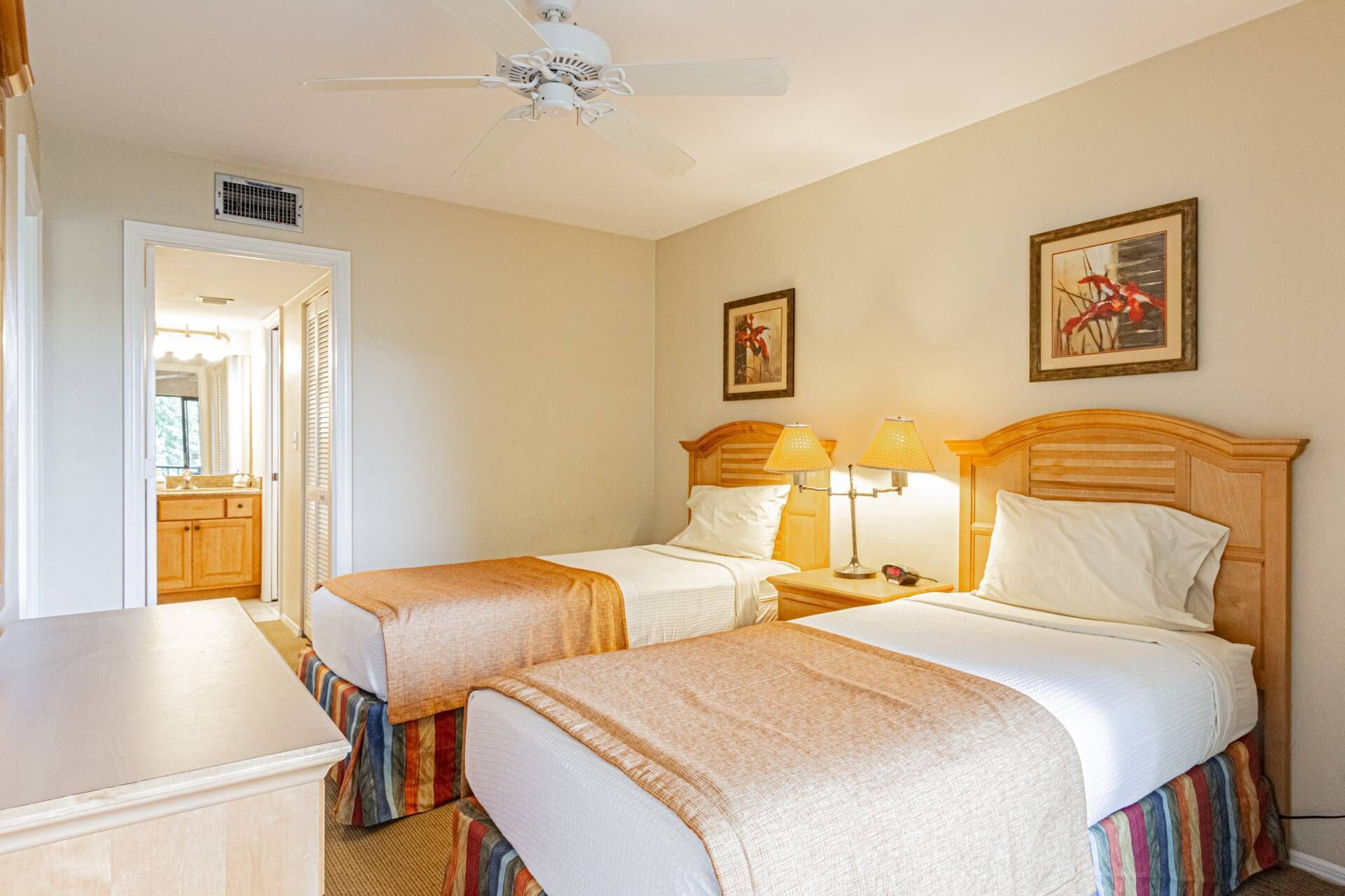 Une chambre d’hôtel avec deux lits, un ventilateur de plafond, une salle de bain avec un meuble-lavabo et deux tableaux encadrés au mur.