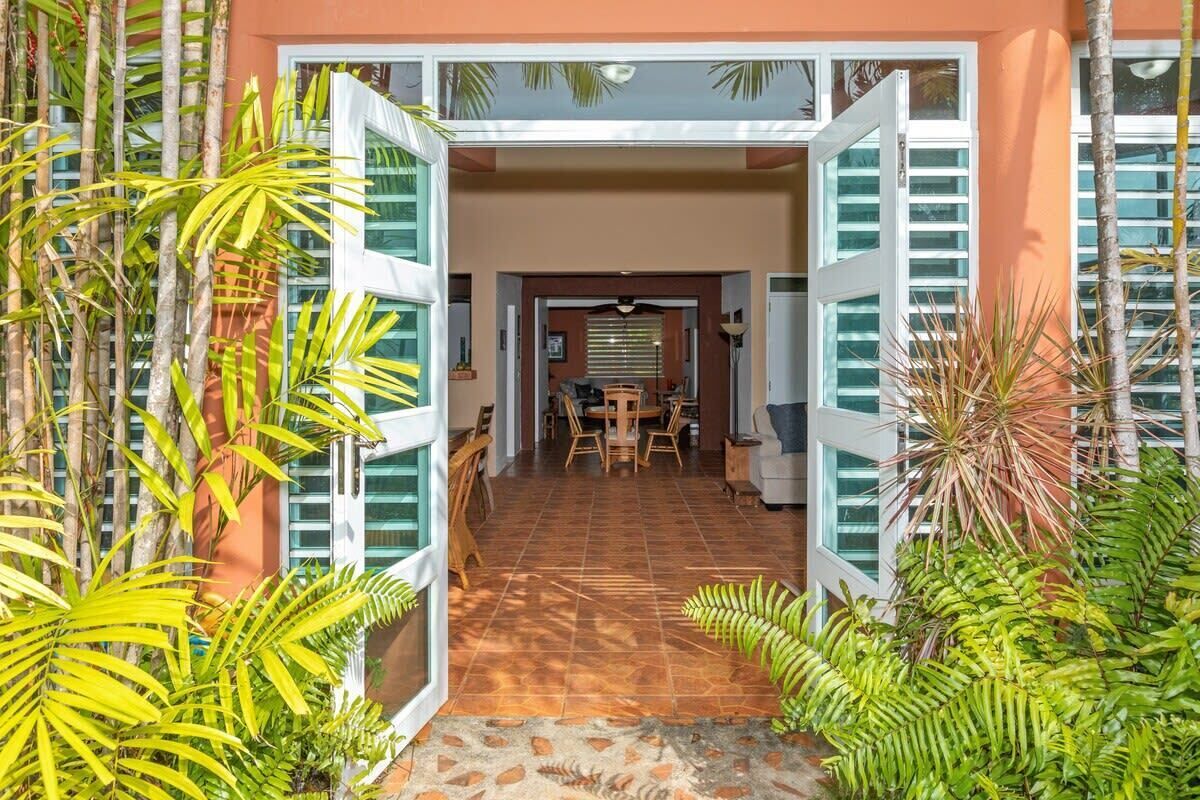 Entrada de hotel con contraventanas abiertas, un área de comedor con mesa y sillas, y plantas tropicales.