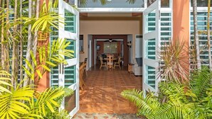 Entrada de hotel com persianas abertas, área de jantar com mesa e cadeiras, e plantas tropicais.