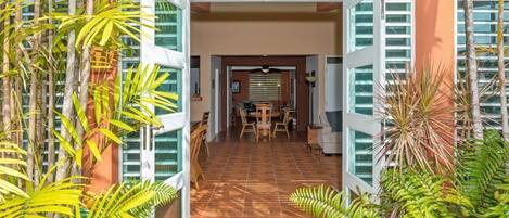 Entrada de hotel con contraventanas abiertas, un área de comedor con mesa y sillas, y plantas tropicales.
