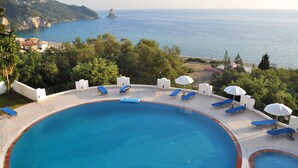Una piscina con sillas de descanso azules y sombrillas, rodeada de vegetación, con vistas a una zona costera y acantilados marinos.