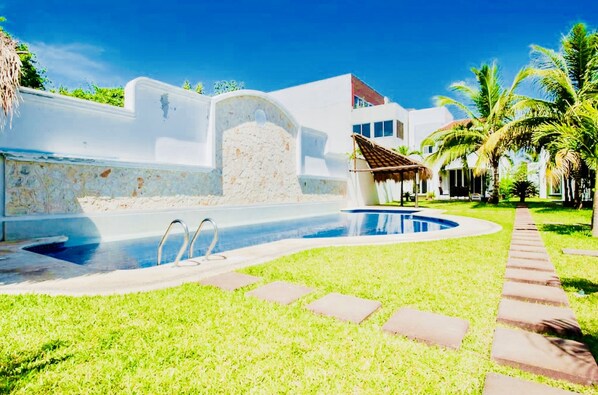 A swimming pool with a stone wall, a thatched-roof structure, and palm trees.