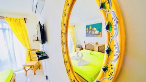 Quarto de hotel com cama verde, uma cadeira de madeira, escrivaninha, televisão e espelho com moldura floral.