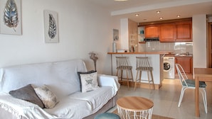 A living room with a sofa, a round coffee table, and a kitchen area with wooden cabinets and a washing machine. - A75 CALPPLACE ESC 1 5º 3 (Calpe)