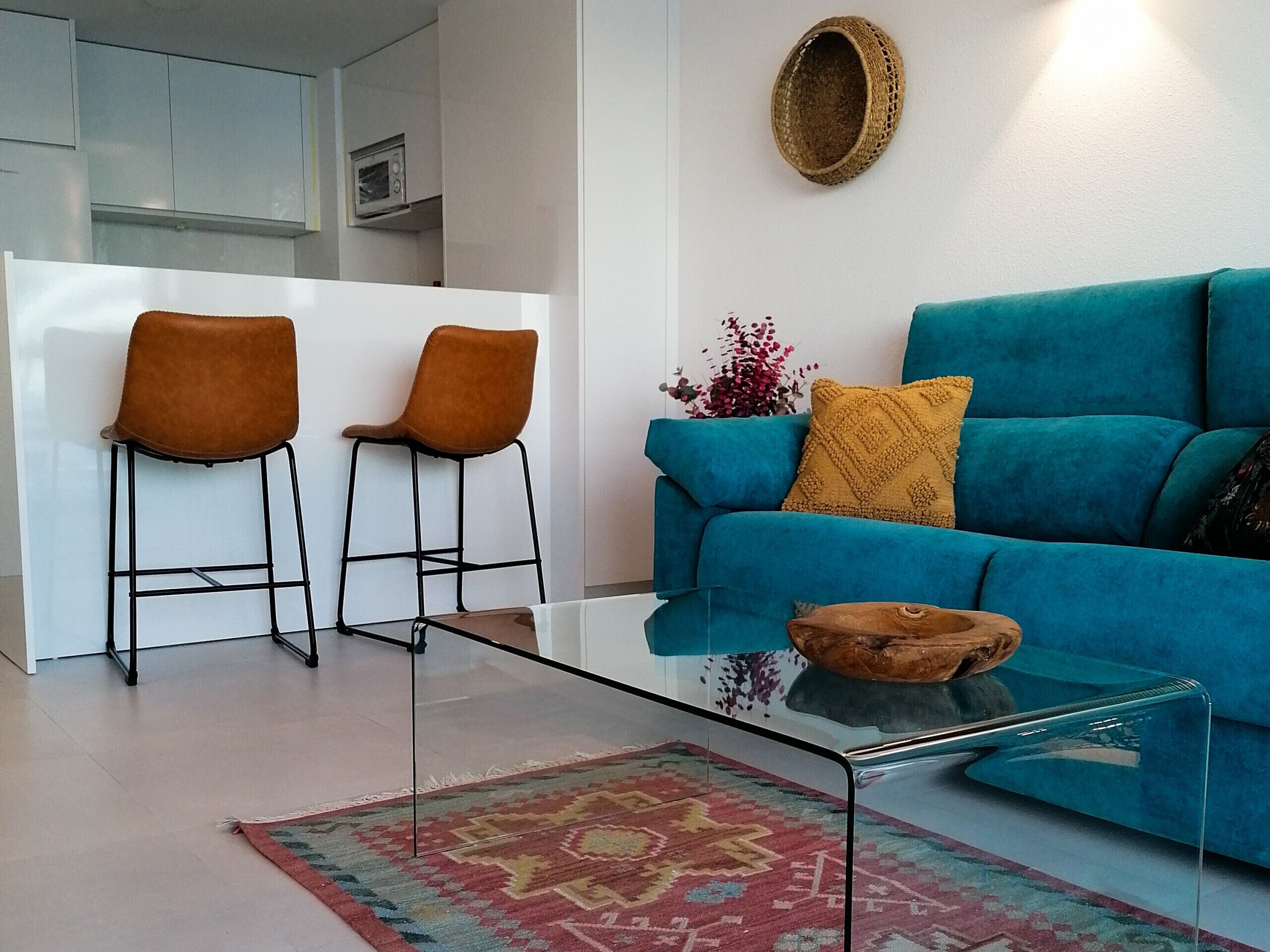 A modern living room with a teal sofa, two brown chairs, a glass coffee table, and a patterned rug.