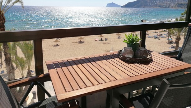 Une table en bois, sur laquelle est posée une plante en pot, se trouve sur un balcon donnant sur une plage parsemée de parasols et fréquentée par des gens.