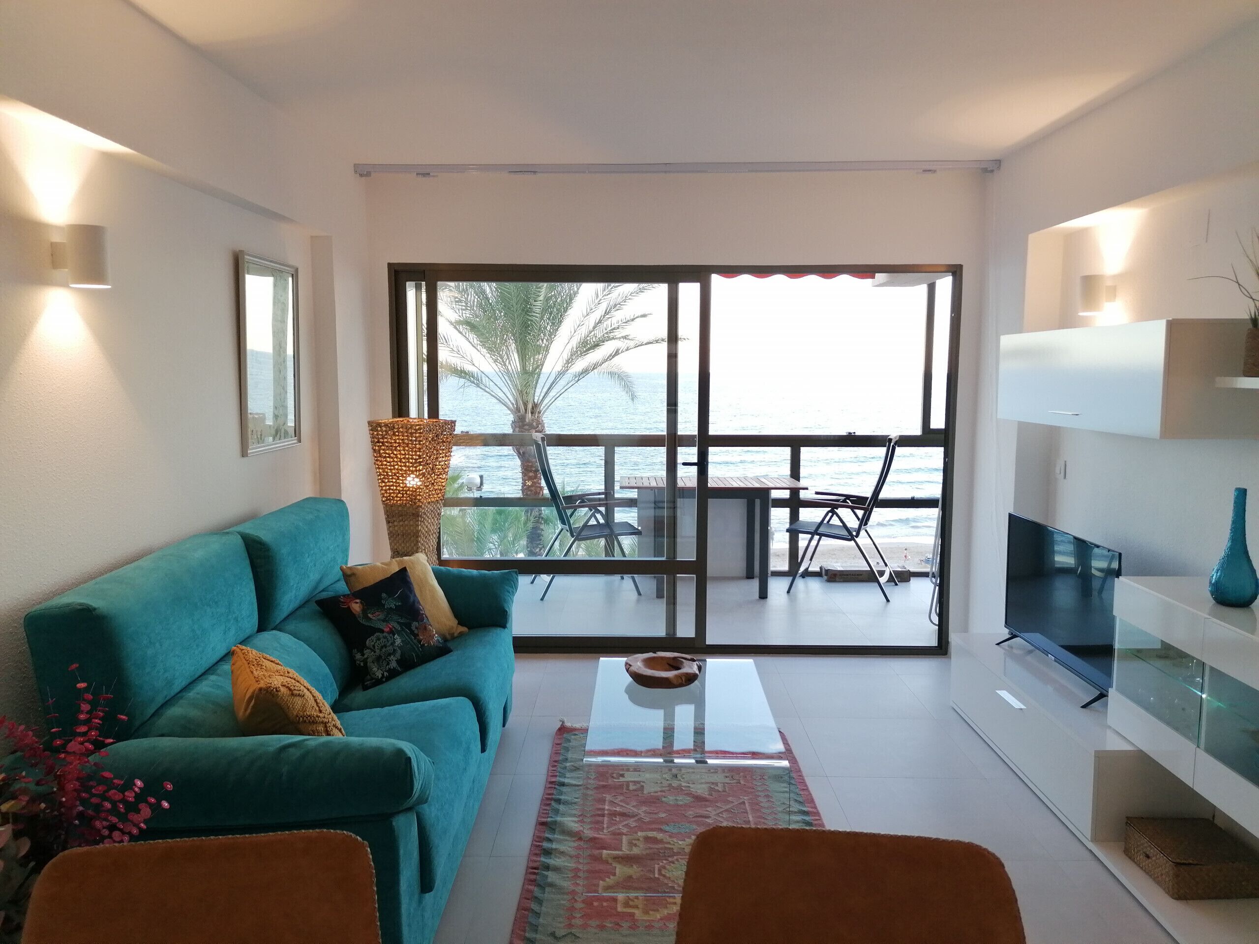 A modern living room with a teal sofa, a glass coffee table, and a view of the sea through large windows.