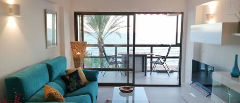 A modern living room with a teal sofa, a glass coffee table, and a view of the sea through large windows.