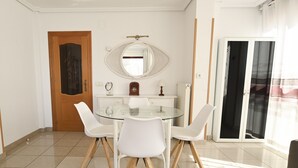 Un'area pranzo con un tavolo in vetro e sedie bianche, una porta in legno, un armadietto bianco e un grande specchio a parete.