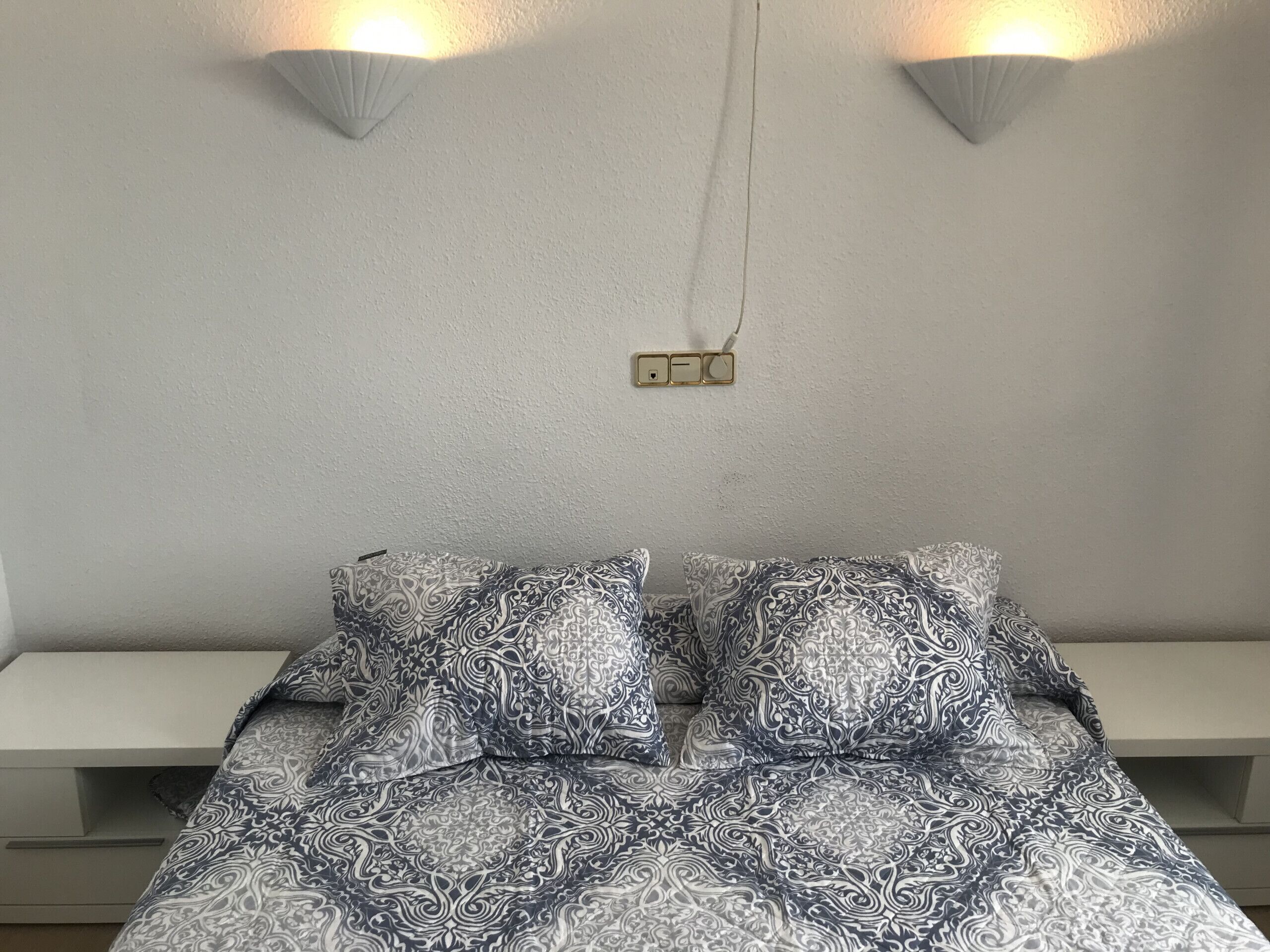 Un dormitorio con una colcha estampada, dos luces fijadas a la pared y un enchufe en la pared.