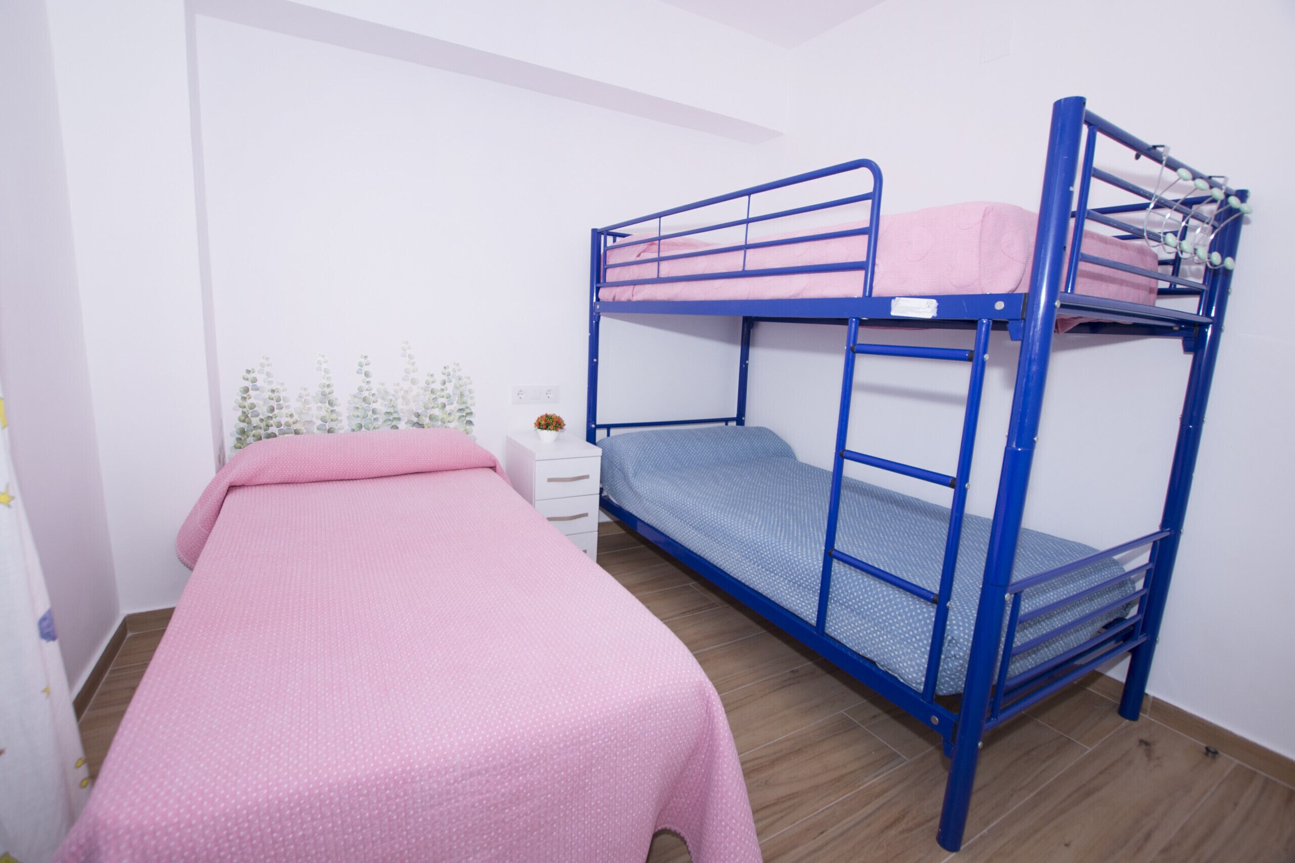 Una habitación con dos literas, una con cubrecama rosa y la otra con cubrecama azul. Hay una mesita de noche blanca con una lámpara y una planta pequeña.