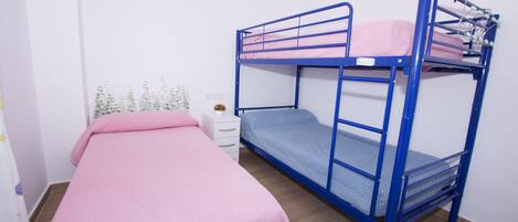 Una habitación con dos literas, una con cubrecama rosa y la otra con cubrecama azul. Hay una mesita de noche blanca con una lámpara y una planta pequeña.