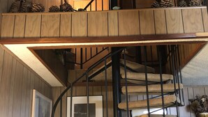Una escalera de caracol con detalles de madera y barandilla, que conduce a un rellano en el segundo piso con piñas y una pequeña mesa.