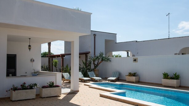 Een zwembadgedeelte met ligbedden en plantenbakken, een bar met een ventilator en een helderblauwe lucht.
