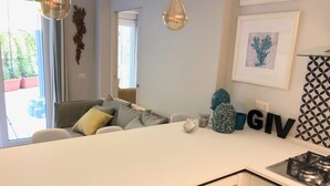 A modern kitchen with a white countertop, a built-in dishwasher, and a gas stove. There is a dining area with a sofa and a coffee table. The room has a large window and a decorative wall hanging. - Casa Mando Terrace With Private Jacuzzi - Taormina (Taormina)