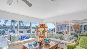 A spacious living room with a large window overlooking a pool and a view of a resort. There is a wooden coffee table with decorative items and a white sofa with colorful cushions.