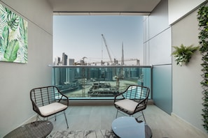 A balcony with two wicker chairs, a glass table, and a view of a cityscape with construction cranes.