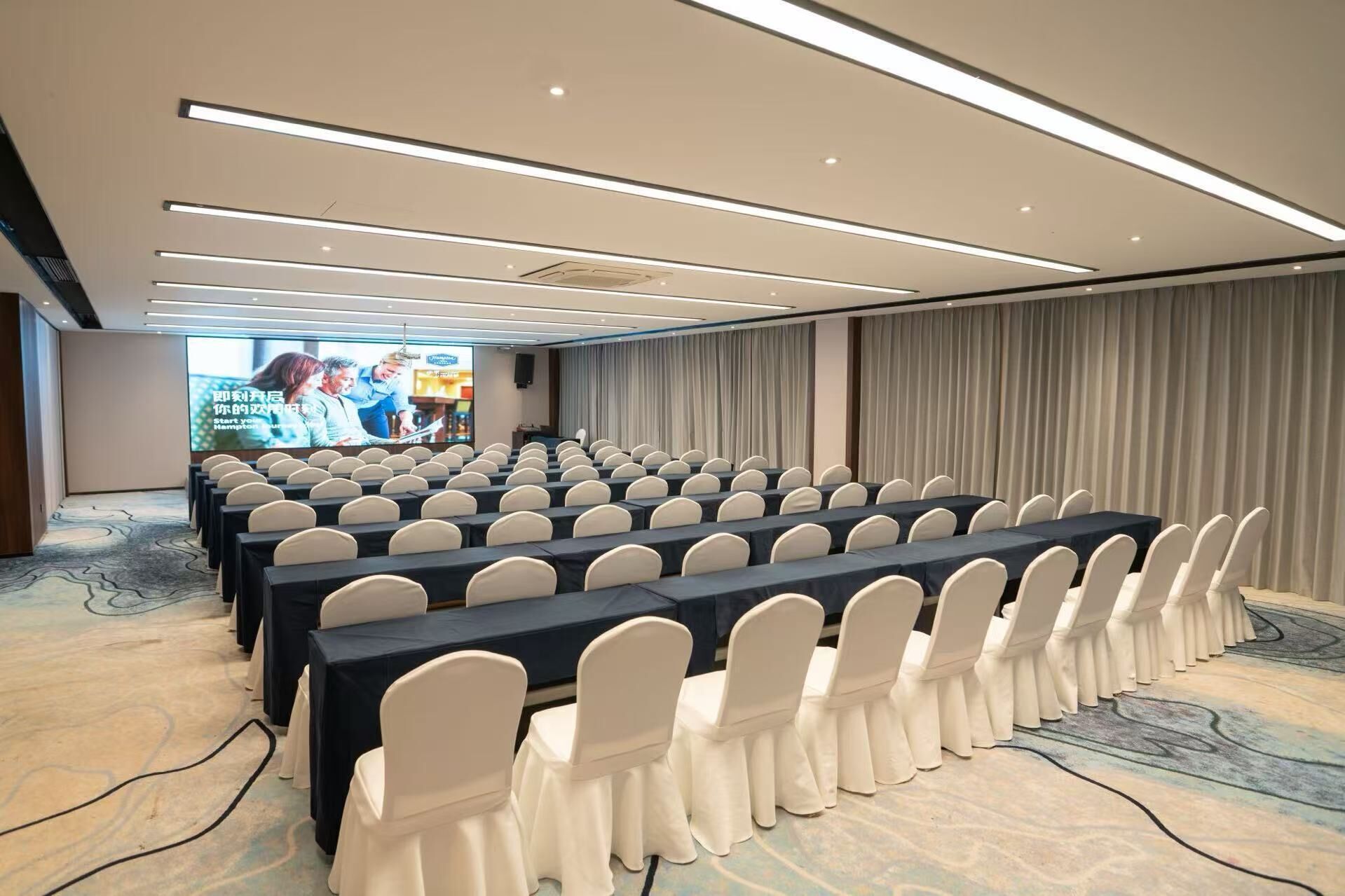 A conference room with rows of tables and chairs, a large screen for presentations, and a carpeted floor.