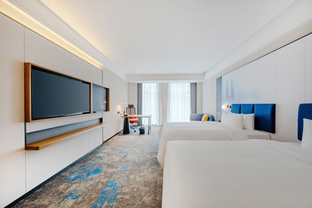 A hotel room with a large flat-screen TV, a desk with a chair, two beds, and a large window with curtains.