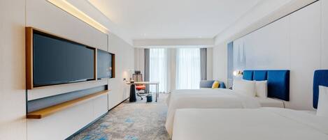 A hotel room with a large flat-screen TV, a desk with a chair, two beds, and a large window with curtains.
