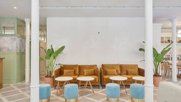 A lobby with a modern design, featuring a central seating area with a sofa and round tables, surrounded by potted plants and white columns.
