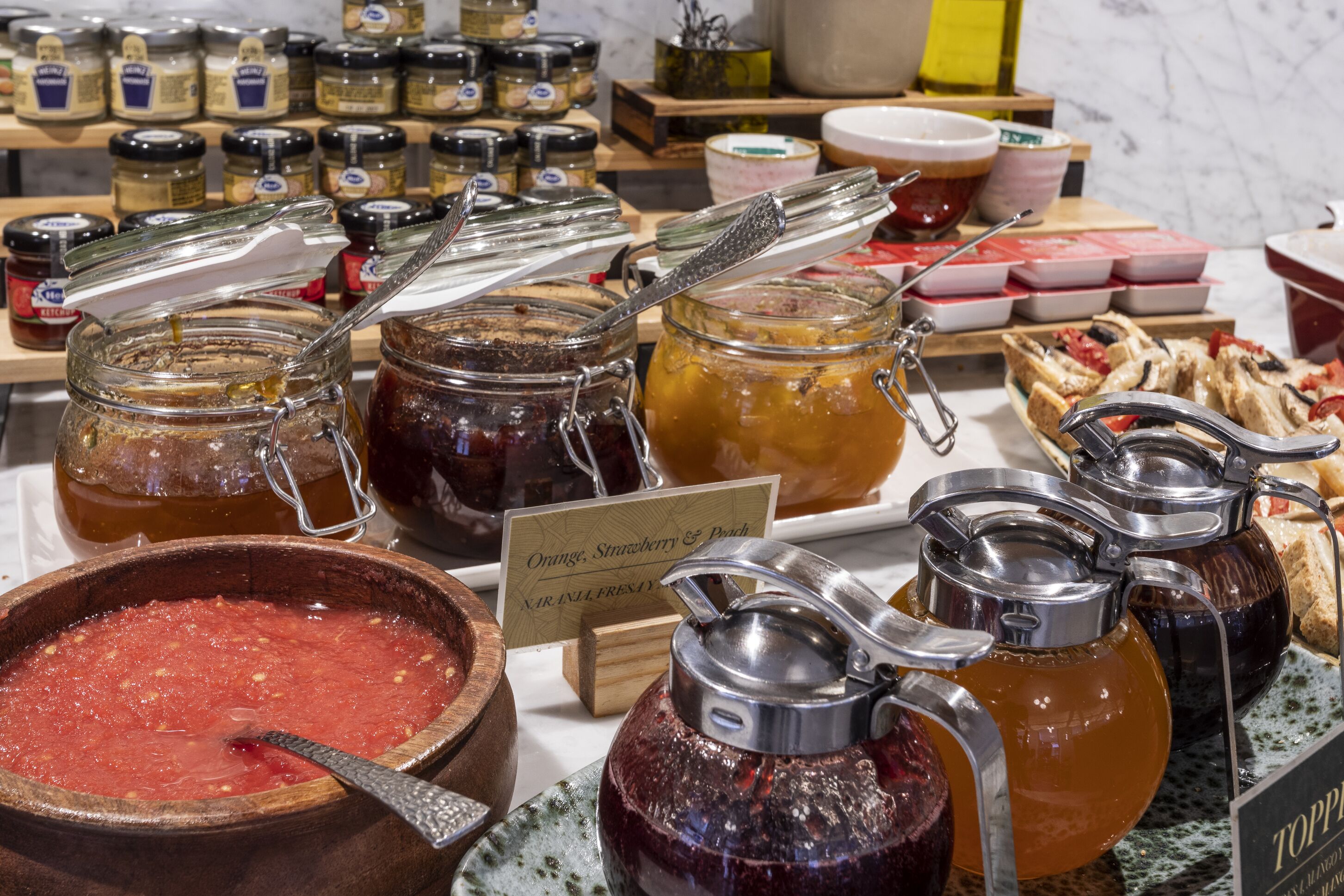 Diversos alimentos, incluyendo frascos de conservas, un cuenco de madera con compota de frutas, y un cartel que indica sabores como naranja, fresa y melocotón.
