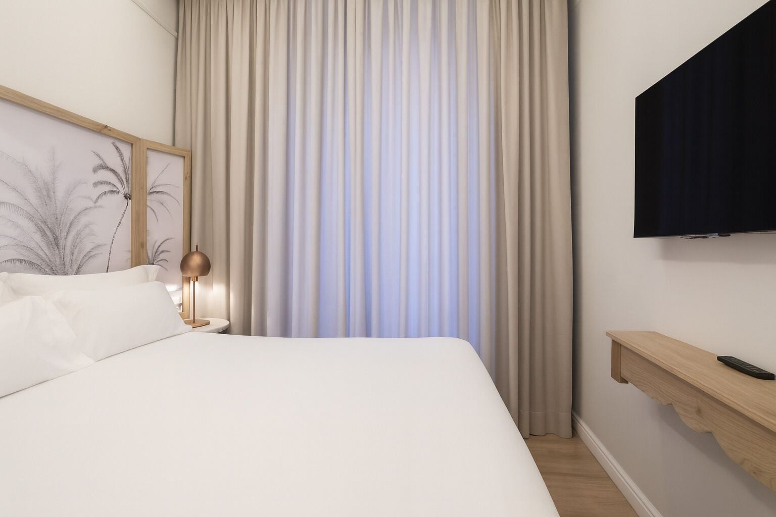 Una habitación de hotel con una cama grande, un televisor montado en la pared, una mesita de noche con lámpara y un estante de madera con control remoto.