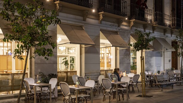 An outdoor cafe with tables and chairs, a tree, and a building with large windows and balconies. - ICON Malabar (Málaga)