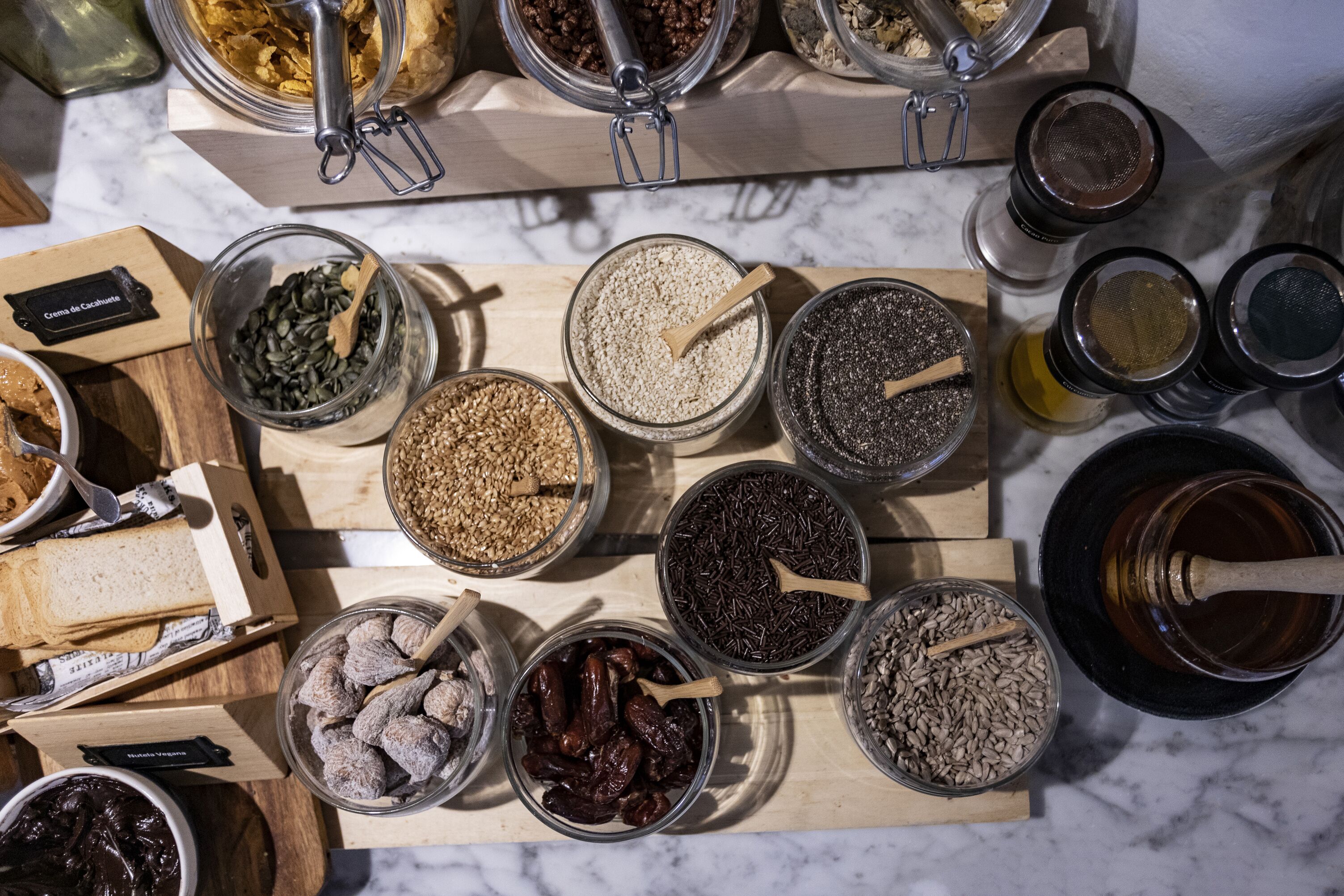 Variedad de alimentos y condimentos dispuestos sobre una tabla de madera, incluyendo frascos con semillas, frutos secos y frutas deshidratadas.
