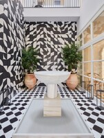 A modern indoor space with a geometric patterned wall, a central white basin, potted plants, and a checkered floor.