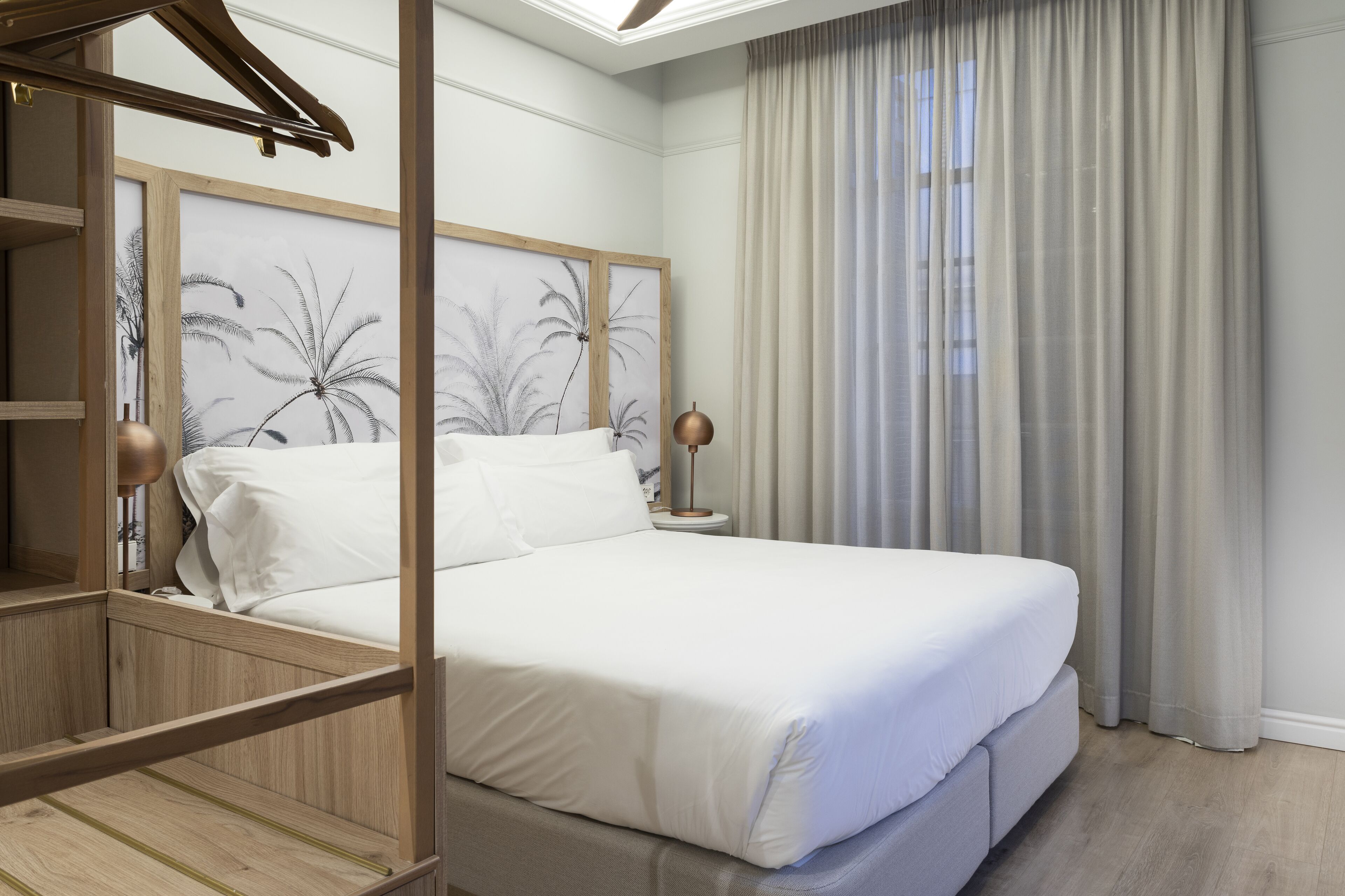 A neatly made bed with white linens in a room with wooden headboard and a large window with sheer curtains.